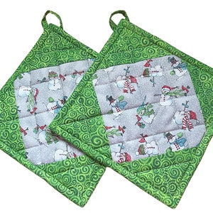 Puede incluir: Dos agarraderas de olla acolchadas de color verde y gris con un patrón de muñeco de nieve. Las agarraderas tienen un patrón de espiral verde en el borde y un centro gris con muñecos de nieve que llevan sombreros y bufandas.