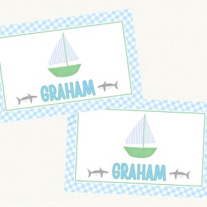 May include: Two blue and white gingham place cards with a green sailboat and two gray sharks. The name "GRAHAM" is printed on each card.
