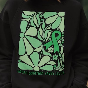 May include: Black sweatshirt with a green floral design and a green ribbon. The text "ORGAN DONATION SAVES LIVES" is printed at the bottom of the design. The sweatshirt is made of a soft material.