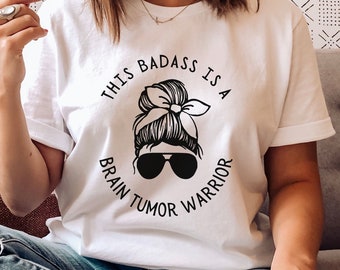 May include: White t-shirt with a black graphic design that says "This badass is a brain tumor warrior". The graphic features a woman with a messy bun and sunglasses.