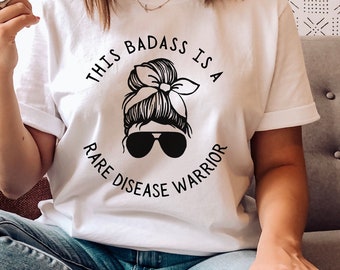 May include: White t-shirt with a black graphic design of a woman with a bun and sunglasses. The text "This badass is a rare disease warrior" is printed on the shirt.