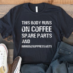 Pode incluir: Uma camiseta preta com texto branco que diz "This body runs on coffee, spare parts and immunosuppressants". A camiseta está colocada sobre uma superfície de madeira com um par de tênis brancos e uma calça jeans azul.