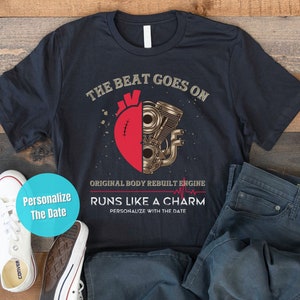 May include: A black t-shirt with a graphic of a heart with a mechanical engine inside. The text "The beat goes on" is above the graphic and "Original body rebuilt engine" is below the graphic. The text "Runs like a charm" is below the engine graphic with the text "Personalize with the date" below that.