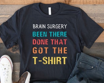May include: A black t-shirt with a vintage-style graphic that reads "Brain Surgery Been There Done That Got The T-Shirt". The shirt is laid out on a wooden surface with a pair of white sneakers and blue jeans.