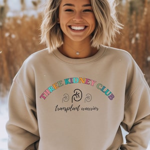 May include: A beige sweatshirt with the text "THREE KIDNEY CLUB" in a rainbow arc with kidney icons and the text "transplant warrior" below.