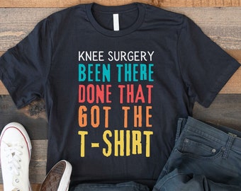 May include: A black t-shirt with the text "Knee Surgery Been There Done That Got The T-Shirt" in rainbow colors. The shirt is laid out on a wooden surface with a pair of white sneakers and blue jeans.