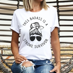 Peut inclure: T-shirt blanc avec un texte noir qui dit "This badass is a stroke survivor". Le texte est en cercle autour d'une illustration en noir et blanc d'une femme avec un chignon désordonné et des lunettes de soleil.