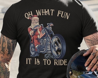 May include: Black t-shirt featuring Santa Claus riding a motorcycle with an American flag design. The shirt has the text "OH WHAT FUN IT IS TO RIDE". The image shows a man with tattoos sitting on a motorcycle.