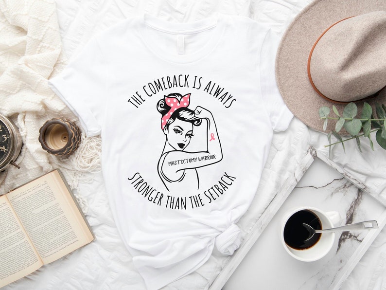 May include: White t-shirt with a black graphic of a woman with a pink polka dot headband and a pink ribbon on her arm. The text reads "The comeback is always stronger than the setback" and "Mastectomy Warrior".