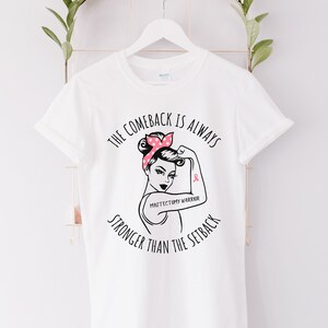 May include: White t-shirt with a black and white illustration of a woman with a pink polka dot bandana and a pink ribbon on her arm. The text "The comeback is always stronger than the setback" is printed on the shirt. The text "Mastectomy Warrior" is printed below the illustration.