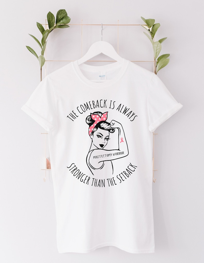 May include: White t-shirt with a black and white illustration of a woman with a pink polka dot bandana and a pink ribbon on her arm. The text "The comeback is always stronger than the setback" is printed on the shirt. The text "Mastectomy Warrior" is printed below the illustration.