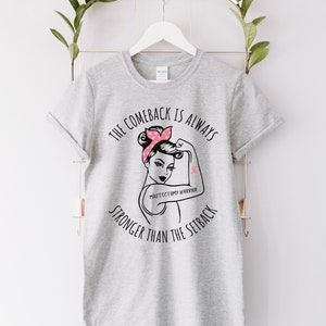 May include: Gray t-shirt with a black graphic of a woman with a pink ribbon on her arm. The text on the shirt reads "The comeback is always stronger than the setback" and "Mastectomy Warrior".