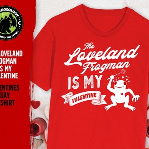 May include: Red t-shirt with a white graphic design featuring a cartoon frog holding a wand with hearts. The text on the shirt reads "The Loveland Frogman is my Valentine".