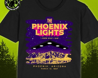 May include: Black t-shirt with a graphic design of a UFO over a cityscape. The text "The Phoenix Lights" is in pink and yellow with the tagline "I know what I saw". The text "Phoenix, Arizona March 13, 1997" is below the graphic.