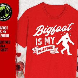 May include: Red t-shirt with a white graphic of Bigfoot walking with a heart on his chest. The text "Bigfoot is my Valentine" is printed on the shirt.