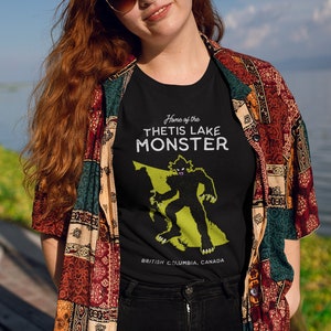 K&ouml;nnte beinhalten: Schwarzes T-Shirt mit einer gr&uuml;nen Silhouette eines Monsters und dem Text "Home of the Thetis Lake Monster" und "British Columbia, Canada".