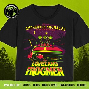 May include: Black t-shirt with a graphic featuring three green frogs on a road, a car, and the text "LOVELAND FROGMEN". The shirt also has the text "AMPHIBIOUS ANOMALIES" and "EXPLORING THE WEIRD".