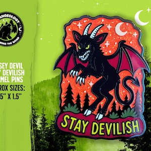 May include: A black enamel pin featuring the Jersey Devil with red wings, horns, and a tail, set against an orange glitter background with pine trees and stars. The text STAY DEVILISH is printed in yellow at the bottom. Measures 1.125 inches by 1.5 inches.