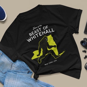 May include: Black t-shirt with a white graphic print of a Bigfoot walking across a yellow outline of the state of New York. The text "Home of the Beast of Whitehall" is printed above the Bigfoot and "New York, USA" is printed below the Bigfoot.