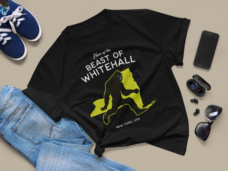 May include: Black t-shirt with a white graphic print of a Bigfoot walking across a yellow outline of the state of New York. The text "Home of the Beast of Whitehall" is printed above the Bigfoot and "New York, USA" is printed below the Bigfoot.