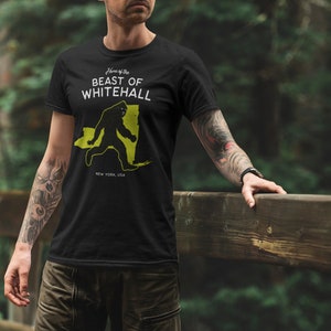 May include: Black t-shirt with a white graphic of a Bigfoot walking across a yellow outline of the state of New York. The text "Home of the Beast of Whitehall" is above the graphic and "New York, USA" is below the graphic.