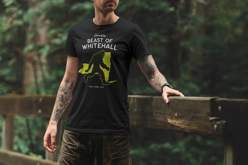 May include: Black t-shirt with a white graphic of a Bigfoot walking across a yellow outline of the state of New York. The text "Home of the Beast of Whitehall" is above the graphic and "New York, USA" is below the graphic.