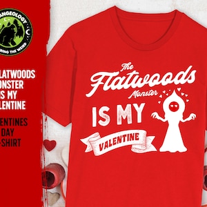 May include: Red t-shirt featuring a white graphic of the Flatwoods Monster, a humanoid figure with a large head and glowing eyes. The text "The Flatwoods Monster is my Valentine" is printed on the shirt.