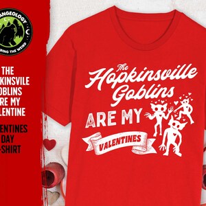 May include: Red t-shirt with white text that reads "The Hopkinsville Goblins Are My Valentines". The text is in a vintage style font. There are three cartoon goblins with hearts above their heads.