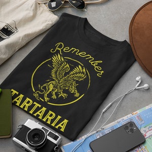 Peut inclure: T-shirt noir avec un graphique jaune d'un lion ail&eacute; avec le texte "Remember Tartaria" dans un cercle.