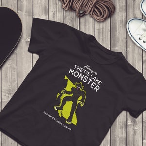 K&ouml;nnte beinhalten: Schwarzes T-Shirt mit einer gr&uuml;nen Grafik eines Monsters auf einer Karte von British Columbia, Kanada. Der Text "Home of the Thetis Lake Monster" ist auf dem Shirt aufgedruckt.