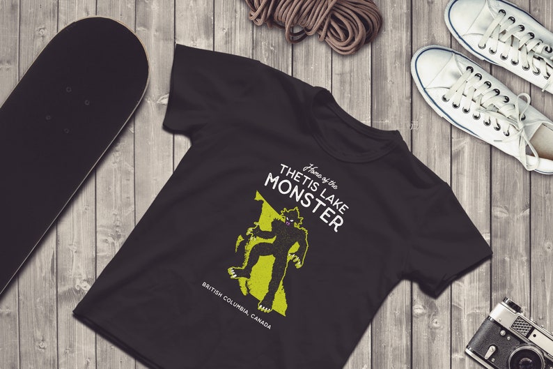 K&ouml;nnte beinhalten: Schwarzes T-Shirt mit einer gr&uuml;nen Grafik eines Monsters auf einer Karte von British Columbia, Kanada. Der Text "Home of the Thetis Lake Monster" ist auf dem Shirt aufgedruckt.
