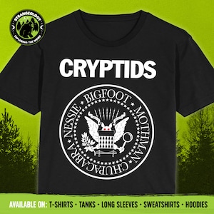 Peut inclure: T-shirt noir avec un graphisme blanc du mot "CRYPTIDS" et un sceau avec les mots "NESSIE BIGFOOT MOTHMAN CHUPACABRA" autour du bord. Le sceau représente une créature stylisée avec des yeux rouges et des ailes.