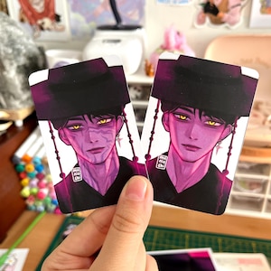 May include: Two rectangular stickers featuring a stylized illustration of a person with purple skin, yellow eyes, and a black hat. The stickers have a dark purple border and the artist's signature.