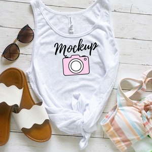 Könnte beinhalten: Ein weißes Tanktop mit einer Kamera-Grafik und dem Wort "Mockup" in schwarzer Schrift. Das Tanktop liegt auf einer weißen Holzoberfläche mit einer braunen Sonnenbrille, weißen und braunen Sandalen und einem gestreiften Badeanzug.