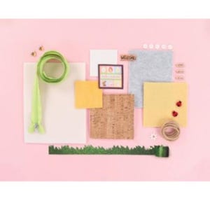 May include: A flat lay of craft supplies on a pink background. Includes a green zipper, felt squares, cork, decorative grass, buttons, and a small framed picture with the word "WELCOME". Decorative elements include ladybugs and bees.