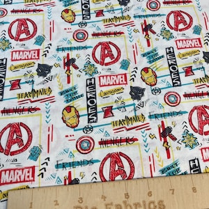 May include: White cotton fabric with a repeating pattern of red, blue, and yellow Marvel superhero characters and logos. The fabric features the Avengers logo, Captain America's shield, Iron Man's helmet, Thor's hammer, Black Panther's mask, and the words "Marvel", "Avengers", "Teammates", "Friends", "Heroes", and "Built for Battle".