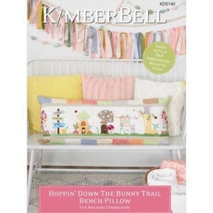 May include: A white bench with a colorful embroidered pillow that says "Hoppin' Down The Bunny Trail Bench Pillow" and "For Machine Embroidery". The pillow features a bunny, mushrooms, and a house.