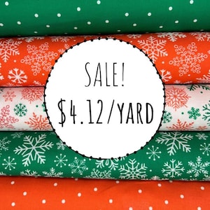 May include: Stacked bolts of fabric featuring festive patterns including green with white polka dots, orange with white snowflakes, white with red and green snowflakes, green with white snowflakes, and orange with white polka dots.