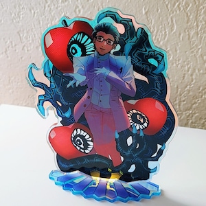 Op de afbeelding: Een kleurrijke acrylstandaard met een personage met roze haar en een wit pak, omringd door zwarte en rode wezens met grote ogen. De standaard heeft een blauwe basis met een wit en geel ontwerp.