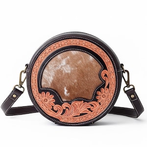 Op de afbeelding: Ronde bruine leren crossbodytas met een koeienhuid middenstuk en bloemmotieven. De tas heeft een afneembare schouderriem en metalen hardware. De diameter van de tas is ongeveer 20 cm.