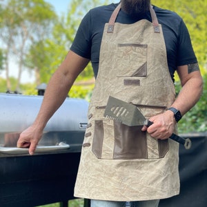 May include: A beige canvas apron with brown leather accents and a large pocket. The apron has adjustable straps and a metal spatula tucked into the pocket.