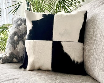 May include: Black and white cowhide pillow with a checkered pattern. The pillow is on a gray couch with a gray and white patterned pillow behind it.