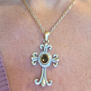 May include: A gold-coloured necklace with a cross pendant. The cross has a central brown gemstone surrounded by small white stones. The cross design has ornate, swirling details. The necklace is worn against a person's skin and a purple top.