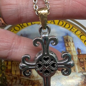May include: Silver pendant necklace with a decorative cross design and heart-shaped cutouts. The cross hangs from a gold-toned bail and a silver and gold chain. The necklace is held against a background with the word "LUCCA".