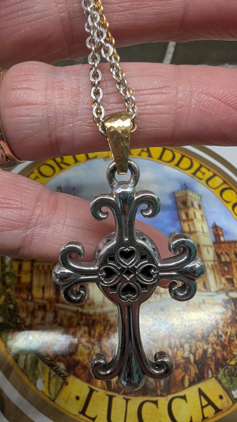 May include: Silver pendant necklace with a decorative cross design and heart-shaped cutouts. The cross hangs from a gold-toned bail and a silver and gold chain. The necklace is held against a background with the word "LUCCA".