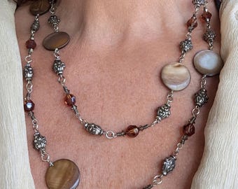 May include: A long, double-strand necklace featuring round, polished brown and cream shell discs interspersed with ornate, textured silver-tone metal beads and small, faceted amber-coloured glass beads connected by silver-tone metal links.