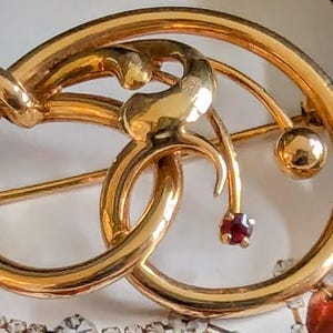 Op de afbeelding: Een gouden broche met een gestileerd bloemmotief en een enkele rode edelsteen. De broche is gemaakt van twee in elkaar grijpende cirkels met een delicaat ontwerp van een stengel en blad.