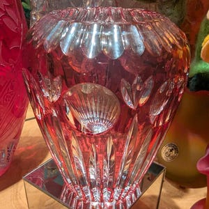 May include: A clear glass vase with a pink hue and intricate cut glass design. The vase has a round body and a wide mouth.