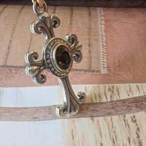 May include: Silver cross pendant with an elaborate design. The cross has a central dark stone set in a gold bezel, encircled by small clear stones. It hangs from a thin, light-brown cord.