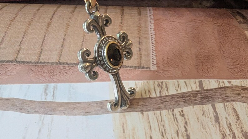 May include: Silver cross pendant with an elaborate design. The cross has a central dark stone set in a gold bezel, encircled by small clear stones. It hangs from a thin, light-brown cord.
